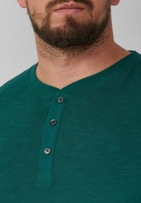 Grünes Kurzarm-Henley-Shirt aus strukturiertem Stoff, mit einem V-Ausschnitt, drei schwarzen Knöpfen und einem schlanken Schnitt.