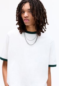 Hvid t-shirt med grøn ribbet krave og manchetter, der har to sølvkæder lagdelt omkring halsudskæringen. Glat stoftekstur.