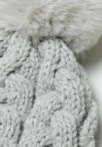 Siva pletena kapa s debelim pletenim uzorkom i lažnim krznom na vrhu, koja prikazuje mekan i teksturiran izgled.