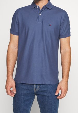 Homme portant un polo bleu à manches courtes avec deux boutons blancs et un jean bleu, debout les mains détendues le long du corps.