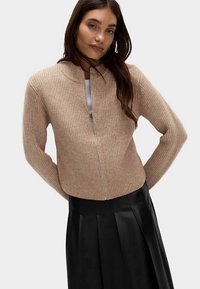 Hellbeiger, gerippter Strickpullover mit durchgehendem Reißverschluss, hohem Kragen, langen Ärmeln und kurzem Schnitt, kombiniert mit einem schwarzen Faltenrock.