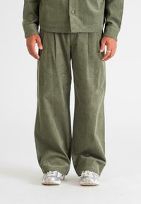 Corduroy outfit in olive green, featuring wide-leg pants with pleats and a button-up shirt. Textured fabric, relaxed fit, paired with gray shoes.
