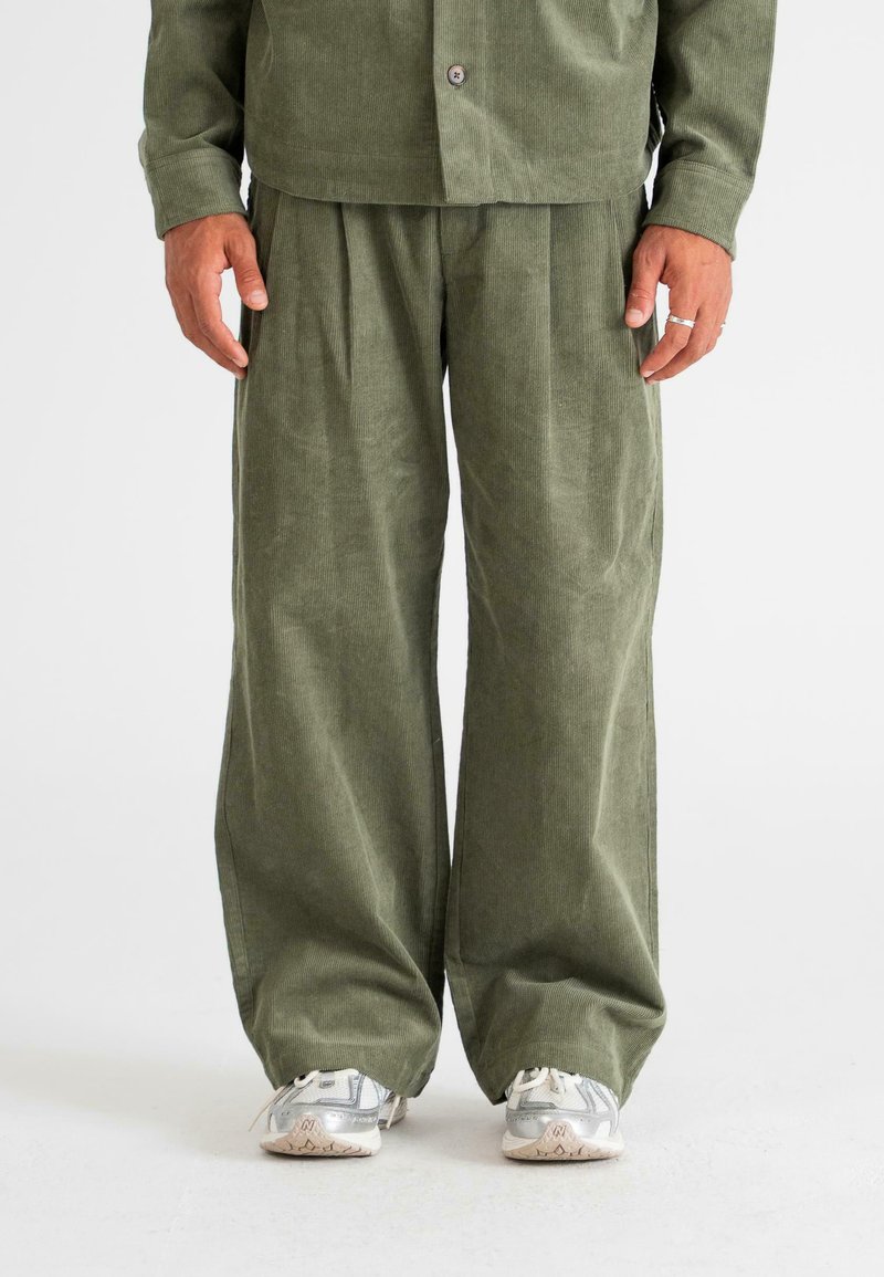 Corduroy outfit in olive green, featuring wide-leg pants with pleats and a button-up shirt. Textured fabric, relaxed fit, paired with gray shoes.
