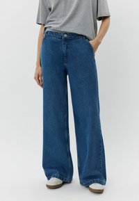 Jeans de denim azul de pierna ancha y cintura alta, con cierre de botón y bolsillos frontales. La tela presenta una textura suave con un color uniforme.