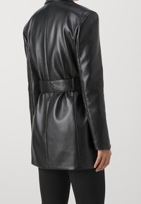 Zwarte leren blazer met een ceintuur in de taille, gladde textuur, gestructureerd ontwerp en een kraag. Beschikt over een getailleerde pasvorm en verticale naden.