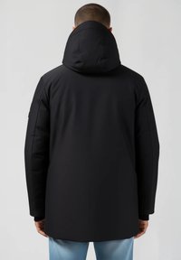 Man i svart huva jacka sedd bakifrån, stående mot en enkel ljusgrå bakgrund.