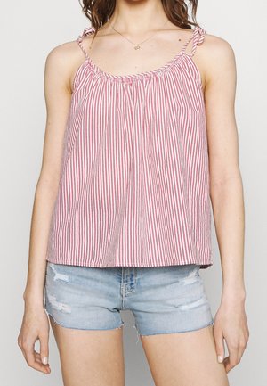 Kobieta ubrana w czerwono-biały, pionowo prążkowany top na ramiączkach z wiązaniami oraz jasnoniebieskie, postrzępione szorty jeansowe.