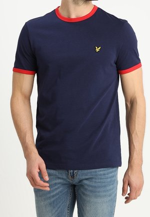 Man draagt een donkerblauw T-shirt met een rode kraag en rode mouwranden, en lichtblauwe spijkerbroek, staand tegen een witte achtergrond.