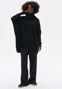 Zwarte oversized trui met een hoge col, geribde manchetten en een zijzak, gecombineerd met zwarte wijde broekspijpen en marineblauwe schoenen.