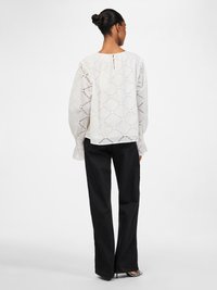 White embroidered blouse featuring a diamond pattern, long sleeves with frill accents, and a back keyhole detail, paired with black wide-leg trousers.