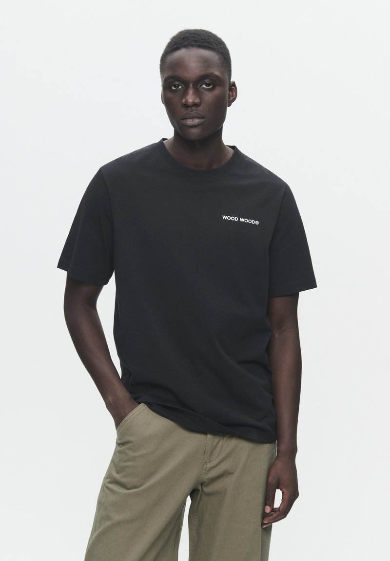 Tricou negru din bumbac cu guler rotund și mâneci scurte, având text alb "WOOD WOOD" pe piept. Asortat cu pantaloni bej.