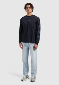 Marineblauw shirt met lange mouwen en logo op de borst en arm, gecombineerd met lichtblauwe spijkerbroek. Model draagt zilveren sneakers. Casual uitstraling.