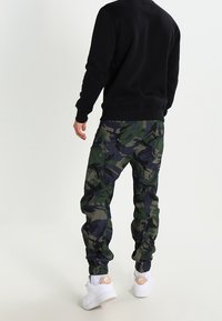 Sudadera negra con pantalones cargo de camuflaje verde y azul marino. Los pantalones cuentan con bolsillos y puños elásticos; combinados con zapatillas blancas.
