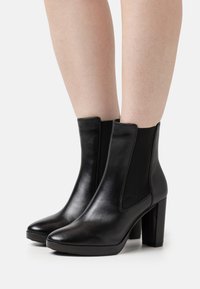Botas pretas até ao tornozelo feitas de couro liso com painéis laterais elásticos, biqueira pontiaguda e um salto grosso. A textura é suave e brilhante.