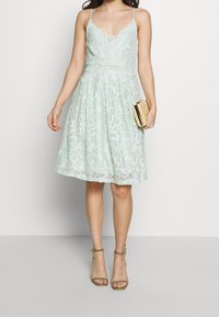 Spetsklänning i mintgrön med V-ringning och scallopade kanter, figursydd ovandel och utsvängd kjol; matchad med en liten kuvertväska och remsandaler.