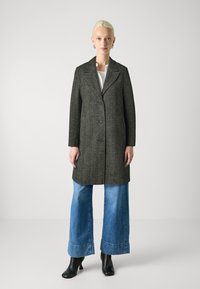 Cappotto grigio texturizzato con colletto a punta, chiusura con bottoni e tasche laterali, indossato sopra jeans denim a gamba larga e stivaletti neri con tacco.