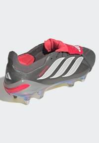 Taco de fútbol Predator con empeine sintético negro, detalles en rojo, franjas plateadas y suela transparente con un toque de rosa.