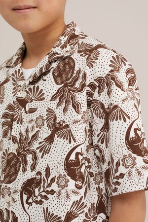 Személy fehér háttéren barna állat- és növénymintás rövid ujjú gombos ingben.