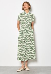 Grün-creamfarbene gemusterte Hemdkleid mit kurzen Ärmeln, Knopfleiste, gebundenem Taillenbund und einem langen, fließenden Rock. Getragen mit hellen Sandalen.
