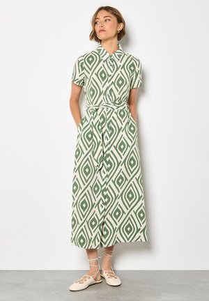 Robe chemise à motifs verts et crème avec des manches courtes, un devant boutonné, une taille à nouer et une jupe longue et fluide. Portée avec des sandales de couleur claire.