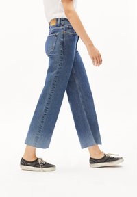 Jeans de tiro alto y pierna ancha en denim azul medio con dobladillo sin acabar. Incluye bolsillos traseros y una etiqueta de cuero marrón en la cintura.