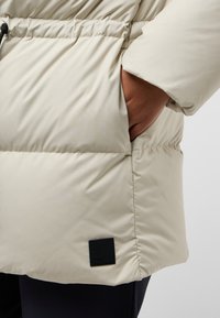 Beige pufferjack met een gestructureerde oppervlakte, elastische tailleband en zijzakken. Heeft een zwart merklabel op de onderrand.