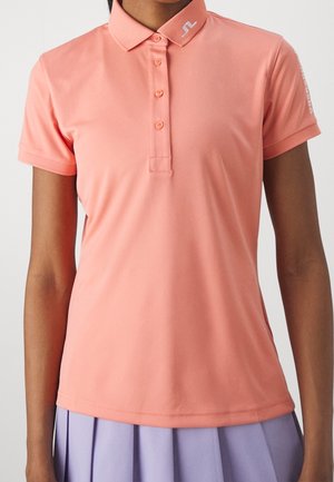 Kvinde iført laksefarvet polo med korte ærmer og knapper samt lavendelfarvet plisseret nederdel, vist fra skuldre til hofter.