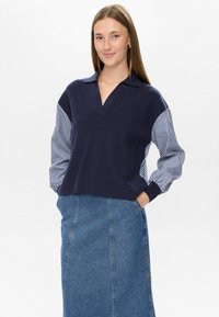 Mlada ženska z dolgimi rjavimi lasmi, oblečena v temno modro jopico z črtastimi rokavi in modrim jeans krilom, stoji s rokami v žepih in se smeška.