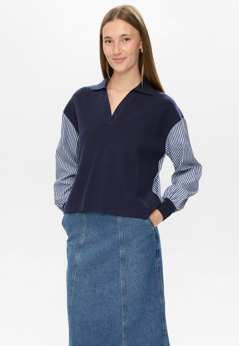 Mlada ženska z dolgimi rjavimi lasmi, oblečena v temno modro jopico z črtastimi rokavi in modrim jeans krilom, stoji s rokami v žepih in se smeška.