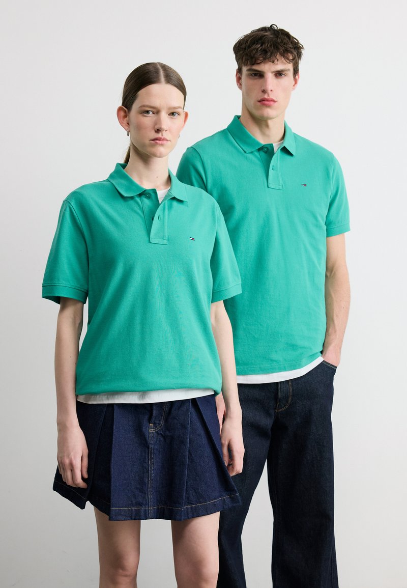 Μια γυναίκα και ένας άντρας στέκονται πλάι-πλάι φορώντας ταιριαστά πετρόλ πόλο μπλουζάκια· η γυναίκα φοράει φούστα από τζιν, ο άντρας σκοτεινό τζιν παντελόνι.