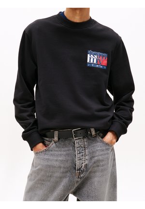 Mann trägt schwarzen Sweatshirt mit blauem, weißem und rotem "Tommy Jeans"-Logo, in hellgraue Jeans mit schwarzem Gürtel gesteckt.
