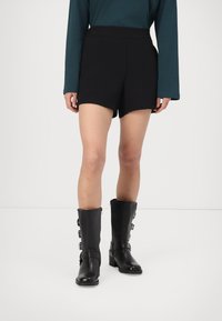 Črne hlače s skrbno oblikovanim krojem imajo rahlo ukrivljen rob. V kombinaciji z zadržnimi črnimi usnjenimi škornji do kolen z zaponkami.