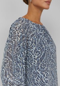 Bluse mit blauem Blumenmuster aus leichtem, transparentem Stoff. Verfügt über einen Rundhalsausschnitt und lange Ärmel. Das Design umfasst verschiedene wirbelnde Formen.