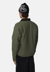 Veste vert olive avec un col en velours côtelé, ornée d'un logo au dos et de poignets boutonnés, associée à un pantalon noir et un bonnet en tricot.