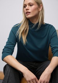 Turquoise gebreide trui met een hoge hals en geribbelde mouwen. Zachte textuur, gecombineerd met donkergrijze broek. Model zit op een houten oppervlak.