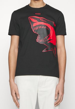T-shirt noir avec un grand graphisme de requin rouge et le texte "HUGO" au-dessus, porté par une personne en pantalon blanc, visible du cou jusqu'au milieu de la cuisse.
