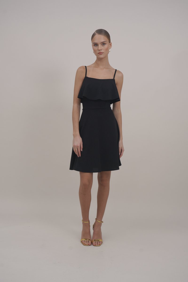 Robe noire avec des bretelles fines, corsage froncé, taille cintrée et jupe évasée. Portée avec des sandales dorées à lanières. Texture de tissu lisse.