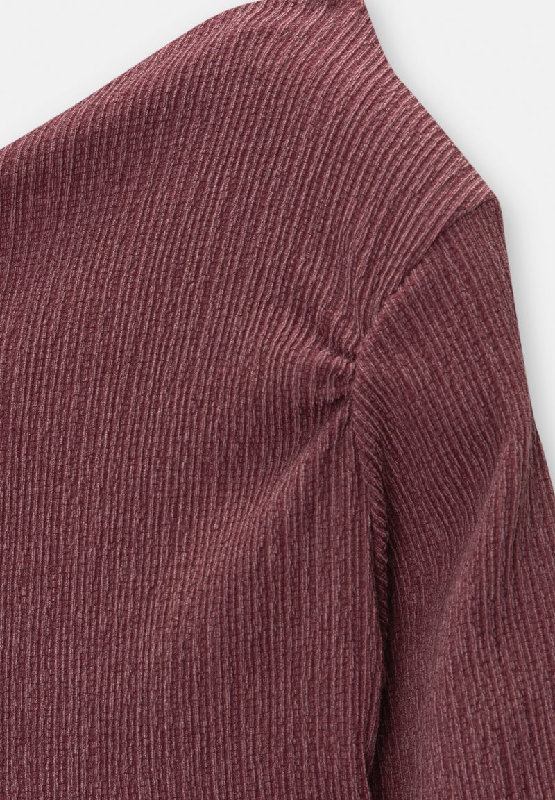 Dettaglio ravvicinato di un tessuto ribbed color bordeaux con cuciture arricciate sulla spalla di un indumento a maniche lunghe.