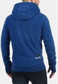Blå fleece-hoodie med hætte, ribbede manchetter og kant. Har et logo på ryggen. Blød tekstur, afslappet design.