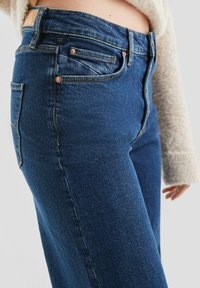 Hoge taille blauwe denim jeans met een gladde textuur, vijf-pocketdesign en koperen knoopaccenten. Close-up zicht dat details en pasvorm toont.
