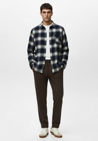 Jeune homme debout, les mains dans les poches, portant une chemise à carreaux bleue marine et blanche par-dessus un tee-shirt blanc, un pantalon marron et des baskets blanches.