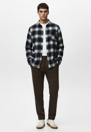 Ung mand stående med hænderne i lommerne iført en marineblå og hvid ternet skjorte over en hvid t-shirt, brune bukser og hvide sneakers.