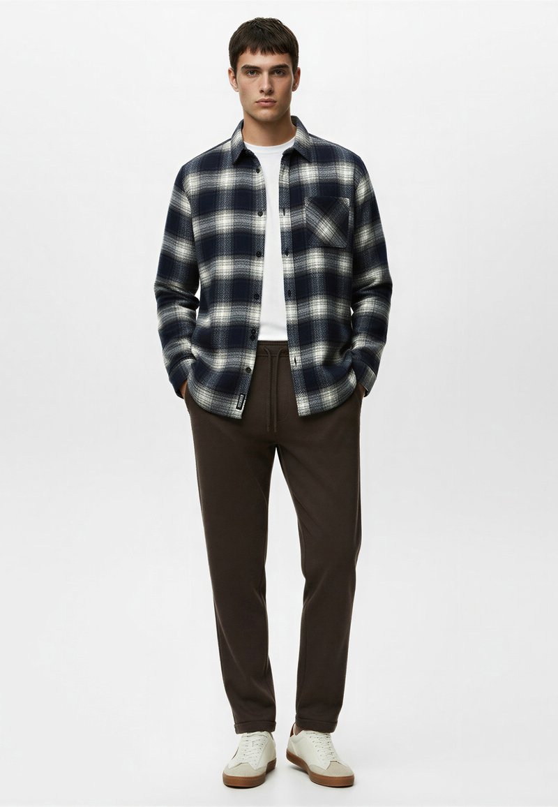 Jeune homme debout, les mains dans les poches, portant une chemise à carreaux bleue marine et blanche par-dessus un tee-shirt blanc, un pantalon marron et des baskets blanches.