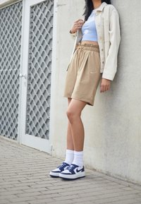 Beiga Nike-shorts med justerbar dragsko, kombinerat med en ljusblå crop top med vita prickar och en krämfärgad jacka. Mörkblå sneakers fulländar looken.