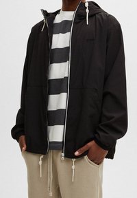 Svart zipjacka med huva, med knytbara dragsko, texturerad tyg och två framfickor. Bärs över en grå och vit randig skjorta.