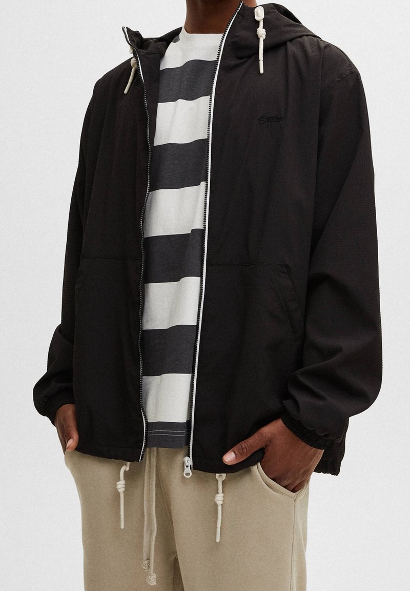 Svart zipjacka med huva, med knytbara dragsko, texturerad tyg och två framfickor. Bärs över en grå och vit randig skjorta.