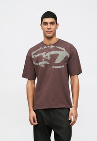 Mladý muž s krátkymi čiernymi vlasmi, oblečený v voľnom hnedom tričku s vyblednutou abstraktnou sivou potlačou a čiernymi nohavicami, stojí pred bielym pozadím.