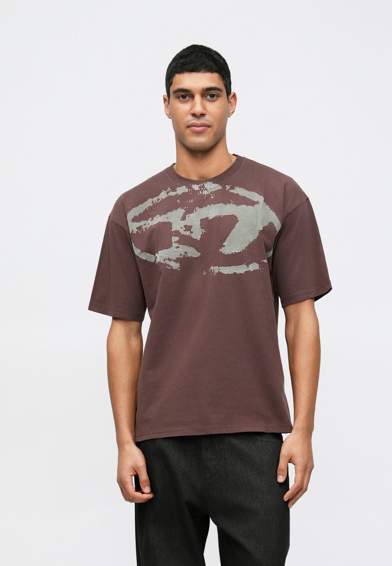 Mladý muž s krátkymi čiernymi vlasmi, oblečený v voľnom hnedom tričku s vyblednutou abstraktnou sivou potlačou a čiernymi nohavicami, stojí pred bielym pozadím.