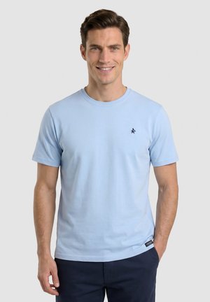 Hombre que lleva camiseta de manga corta azul claro con pequeño logo oscuro en el pecho y pantalones oscuros, sonriendo y de pie frente a un fondo liso.