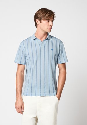 Homme portant un polo à manches courtes bleu clair à rayures verticales et un pantalon blanc, debout avec une main dans la poche, regardant sur le côté.